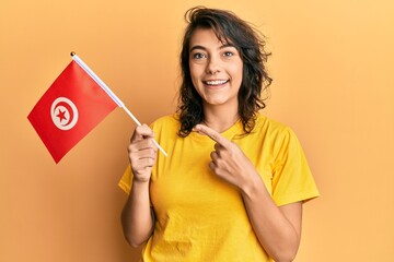 Young hispanic woman holding tunisia flag smiling happy pointing with hand and finger