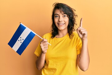 Young hispanic woman holding honduras flag smiling with an idea or question pointing finger with...