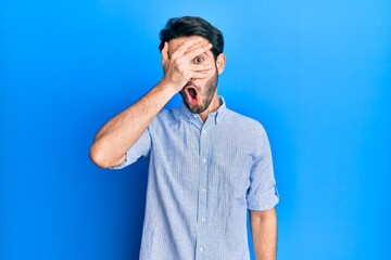 Young hispanic man wearing elegant summer shirt peeking in shock covering face and eyes with hand, looking through fingers afraid