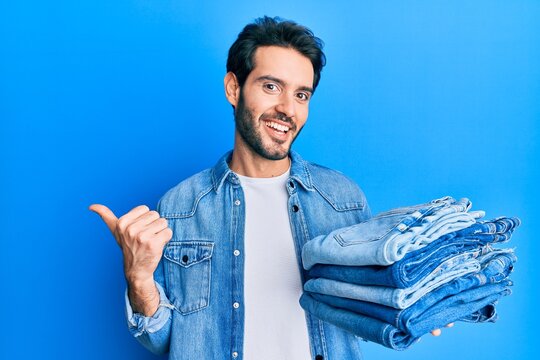 Young hispanic man holding stack of folded jeans pointing thumb up to the side smiling happy with open mouth