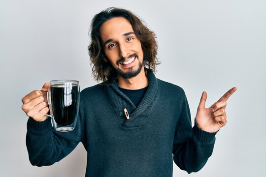 Young hispanic man drinking a cup of coffee smiling happy pointing with hand and finger to the side