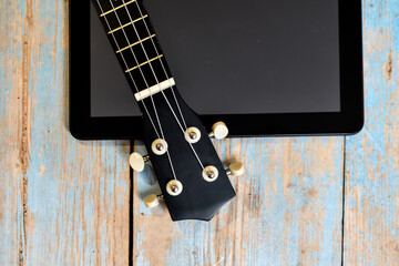 Fototapeta premium Tablet and ukulele on wooden table with space for text. Online learning to play Hawaiian guitar. ukulele fretboard on textured wooden background next to tablet screen