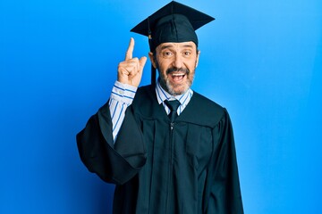 Middle age hispanic man wearing graduation cap and ceremony robe pointing finger up with successful idea. exited and happy. number one.