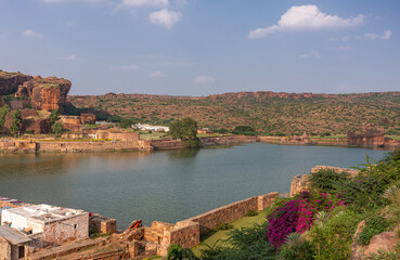 Fototapeta na wymiar Badami, Karnataka, India - November 7, 2013: Cave temples above Agasthya Lake in NE-view, surrounded by hills, rocks, and cliffs under blue cloudscape. Flowers add color, Green foliage.