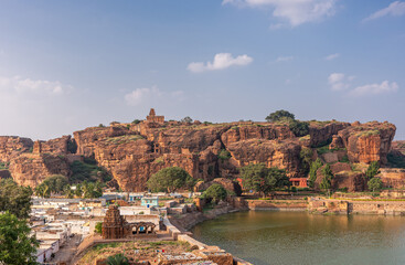 Badami, Karnataka, India - November 7, 2013: Cave temples above Agasthya Lake. Yallamma Temple in front, Upper Shivalaya Temple on top of red rock cliffs. Cityscape at bottom left.