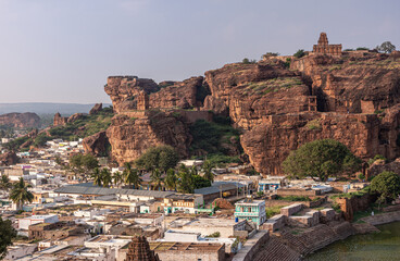 Fototapeta na wymiar Badami, Karnataka, India - November 7, 2013: Cave temples above Agasthya Lake. Cityscape in front of red rock cliffs with Upper Shivalaya temple on top under blue cloudscape,