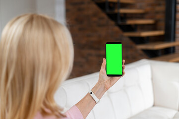 Back view a woman holding smartphone with an empty green screen, a woman sits on the couch at home and holds gadget with blank display, mock-up
