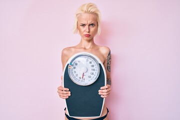 Young blonde woman with tattoo standing shirtless holding weighing machine skeptic and nervous, frowning upset because of problem. negative person.