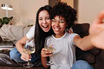 Selfie made on smartphone. Beautiful girls dressed in jeans and bright T-shirts with beautiful smiles and cute make-up drink wine and have good rest