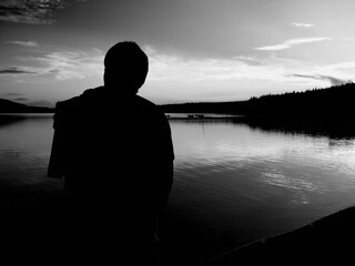 Fototapeta premium Black and white silhouette of a person on lake Loch Lomond during sunset with some clouds and a background forest