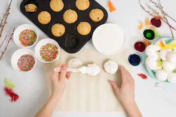 the process of decorating mini cupcakes Easter cakes with white icing and sweet candies, top view, willow branches and eggs for coloring