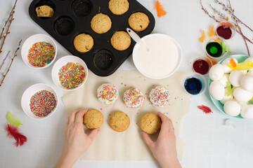 the process of decorating mini cupcakes Easter cakes with white icing and sweet candies, top view, willow branches and eggs for coloring