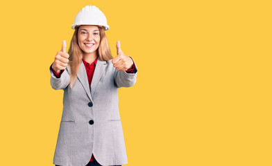 Beautiful young woman wearing architect hardhat approving doing positive gesture with hand, thumbs up smiling and happy for success. winner gesture.
