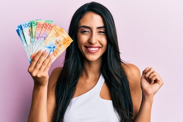 Beautiful hispanic woman holding swiss franc banknotes screaming proud, celebrating victory and success very excited with raised arm