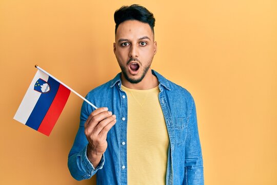Young arab man holding slovenia flag scared and amazed with open mouth for surprise, disbelief face