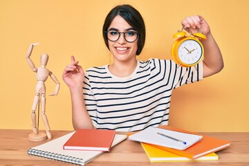 Brunette teenager girl holding alarm clock studying for exam smiling happy pointing with hand and finger to the side