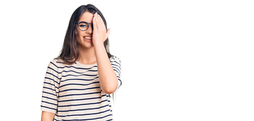Brunette teenager girl wearing casual clothes and glasses covering one eye with hand, confident smile on face and surprise emotion.