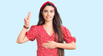Brunette teenager girl wearing summer dress smiling swearing with hand on chest and fingers up,...