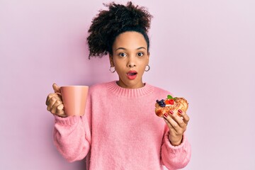 Young african american girl drinking a cup of coffee and eating sweet afraid and shocked with...