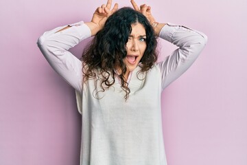 Naklejka premium Young brunette woman with curly hair wearing casual clothes posing funny and crazy with fingers on head as bunny ears, smiling cheerful