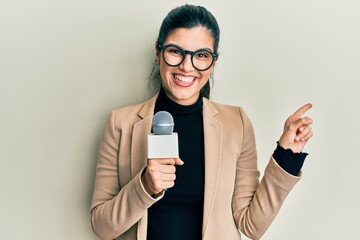 Young hispanic woman holding reporter microphone smiling happy pointing with hand and finger to the...