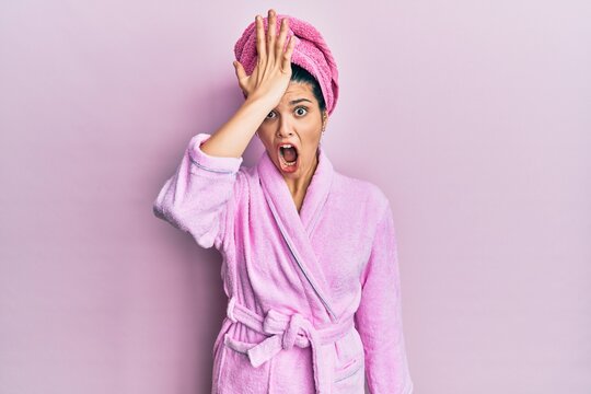 Young hispanic woman wearing shower towel cap and bathrobe surprised with hand on head for mistake, remember error. forgot, bad memory concept.