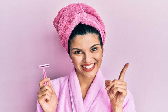 Young hispanic woman wearing shower bathrobe holding razor smiling happy pointing with hand and finger to the side
