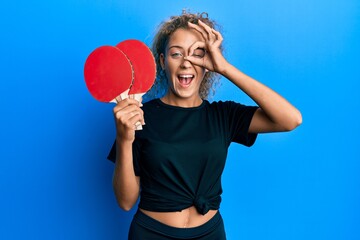 Beautiful caucasian teenager girl holding red ping pong rackets smiling happy doing ok sign with...