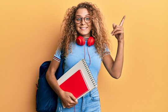 Beautiful caucasian teenager girl wearing student backpack and holding books surprised with an idea or question pointing finger with happy face, number one