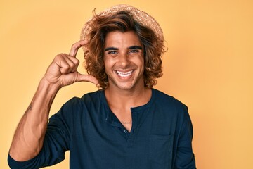 Young hispanic man wearing summer hat smiling and confident gesturing with hand doing small size...
