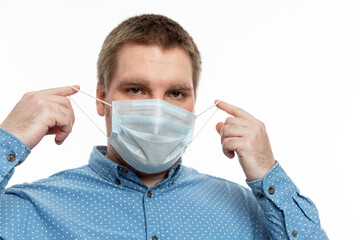 A young man in a blue shirt puts on a medical mask. Quarantine during the coronavirus pandemic. White background.