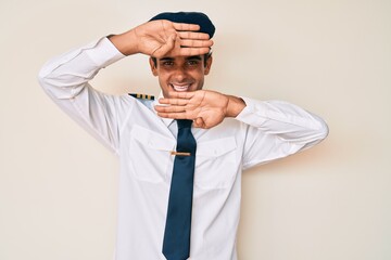 Young hispanic man wearing airplane pilot uniform smiling cheerful playing peek a boo with hands showing face. surprised and exited