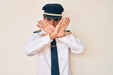 Young hispanic man wearing airplane pilot uniform rejection expression crossing arms doing negative sign, angry face