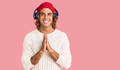 Young hispanic man listening to music using headphones praying with hands together asking for forgiveness smiling confident.