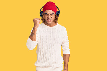 Fototapeta premium Young hispanic man listening to music using headphones angry and mad raising fist frustrated and furious while shouting with anger. rage and aggressive concept.