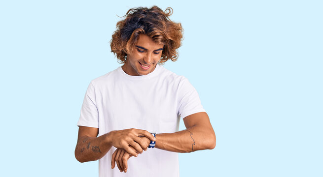 Young hispanic man wearing casual white tshirt checking the time on wrist watch, relaxed and confident
