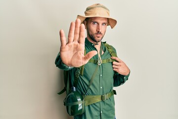 Handsome man with beard wearing explorer hat and backpack doing stop sing with palm of the hand....