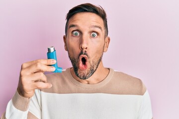 Handsome man with beard holding medical asthma inhaler scared and amazed with open mouth for...