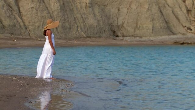 Woman alone in a white dress and hat standing in front of the sea on a deserted beach thinking, nostalgia, emotions, memories 