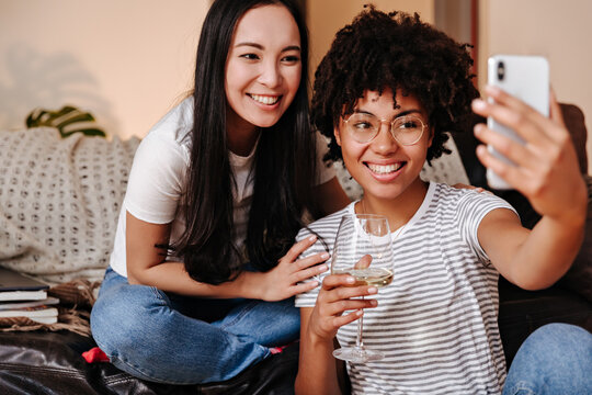 Portrait photo in casual homely setting. Two girlfriends have rest surrounded by books and make cool selfies
