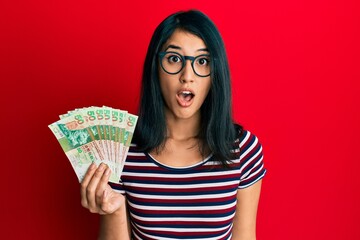 Beautiful asian young woman holding 50 hong kong dollars banknotes scared and amazed with open mouth for surprise, disbelief face