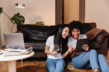Obraz premium Cute girls look at tablet and laugh. Women in jeans sit on floor, leaning on sofa