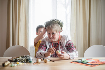 Obraz na płótnie Canvas Retired woman painted easter egg in Ukrainian shirt. DIY craft easter eggs. Grandson put on the head willow wreath his grandmother