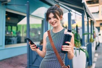 Young caucasian businesswoman using smartphone holding bottle of water at the city.