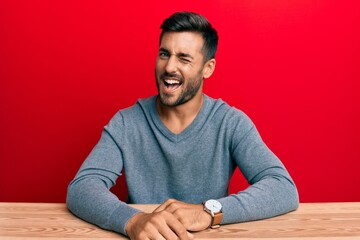 Handsome hispanic man wearing casual style sitting on the table winking looking at the camera with...