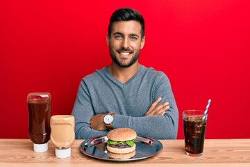 Handsome hispanic man eating a tasty classic burger and soda happy face smiling with crossed arms...