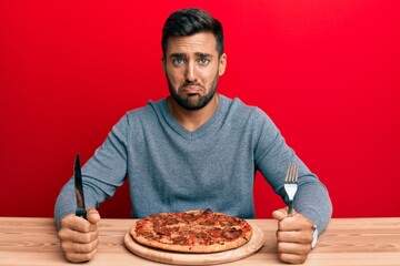 Handsome hispanic man eating tasty pepperoni pizza depressed and worry for distress, crying angry and afraid. sad expression.