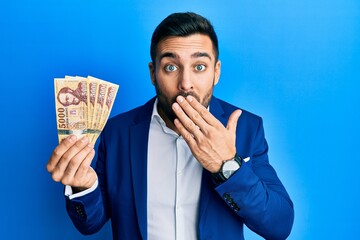 Young hispanic businessman wearing business suit holding hungarian forint banknotes covering mouth...