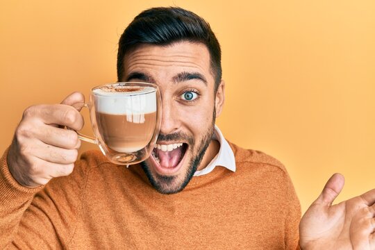 Young hispanic man holding cup of coffee over eye celebrating achievement with happy smile and winner expression with raised hand