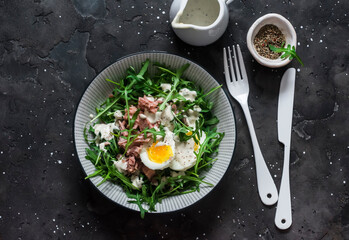 Salad with arugula, canned tuna, boiled egg and yogurt, mustard dressing on a dark background, top view